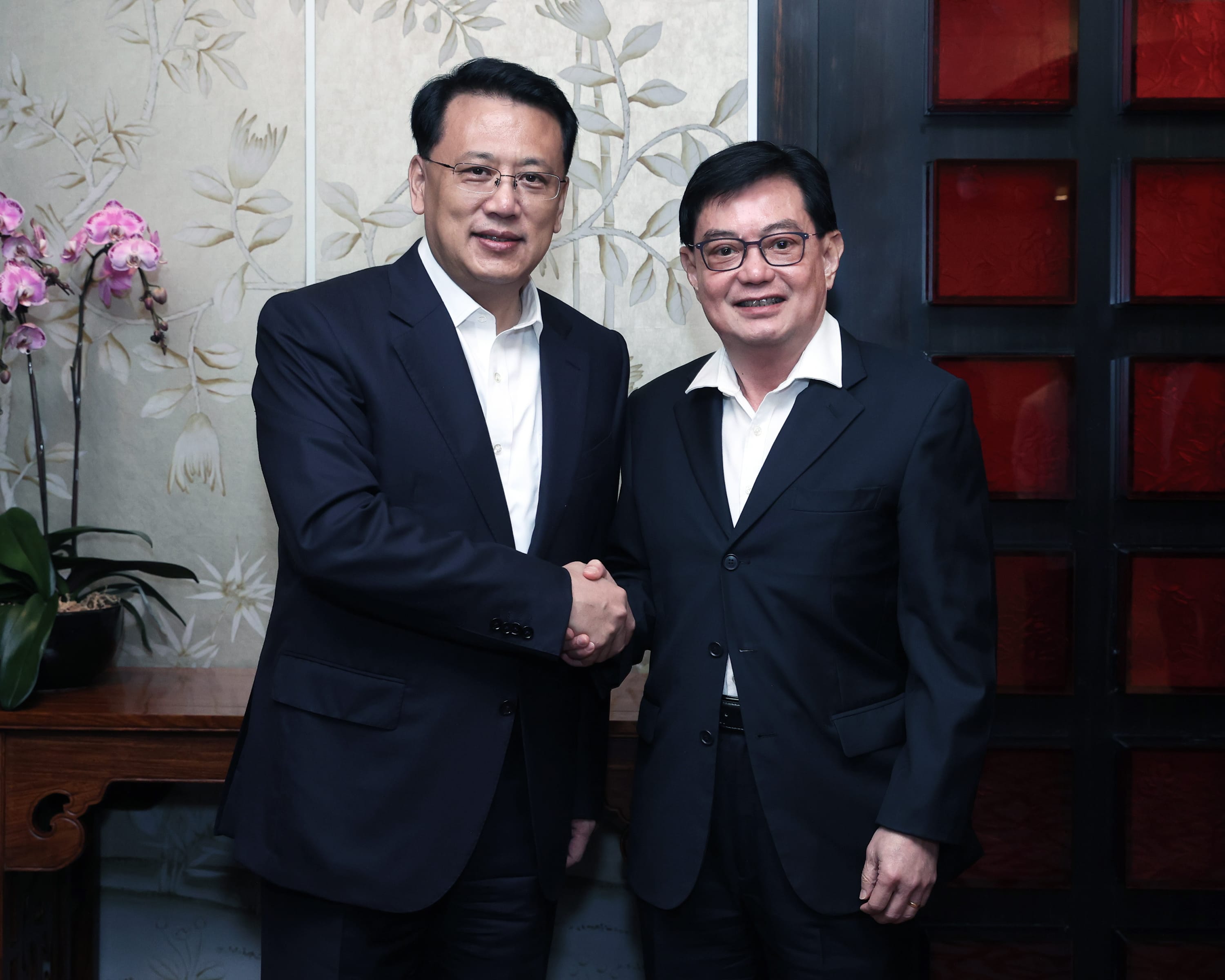 Two men in suits shake hands in front of a floral wall and red-paneled door.